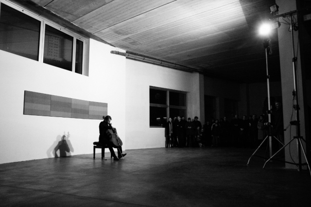 Un violoncellista seduto suona nella sala della mostra. Dietro di lui è appesa un'opera formata da quattro tele accostate in orizzontale. Sulla destra in piedi si vede il pubblico in ascolto. La foto è in bianco e nero.