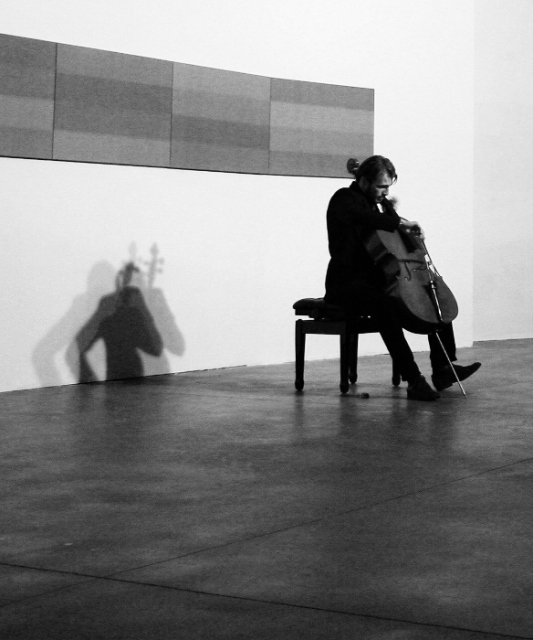 Un violoncellista seduto suona nella sala della mostra. Dietro di lui è appesa un'opera formata da quattro tele accostate in orizzontale. La foto è in bianco e nero.
